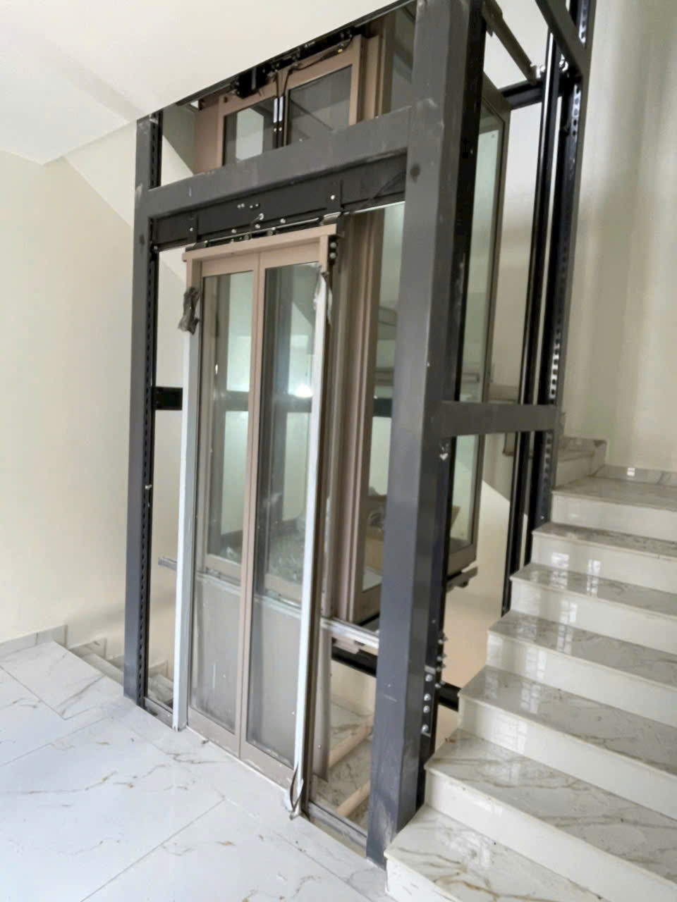 Thang máy gia đình khung kính đang lắp đặt cạnh cầu thang đá trong nhà